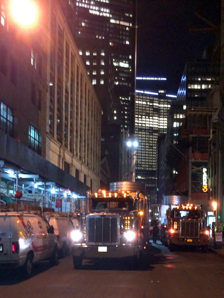 Two trucks towing flatbed with cooling tower during the night with New York City skyline on the background